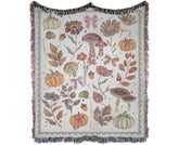 Light cream 100% cotton woven throw blanket featuring mushrooms, autumn leaves, pumpkins with bows, and fringed edges.