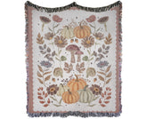 A cotton woven throw blanket with fringe edges, featuring an autumn-themed pattern of pumpkins, mushrooms, leaves, and flowers in muted orange, brown, beige, and gray tones on a cream background.